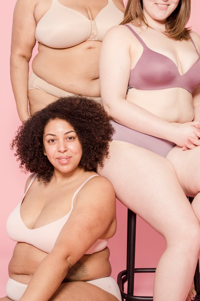 Cheerful plump African American woman in underwear looking at camera against crop unrecognizable multiethnic partners on pink background